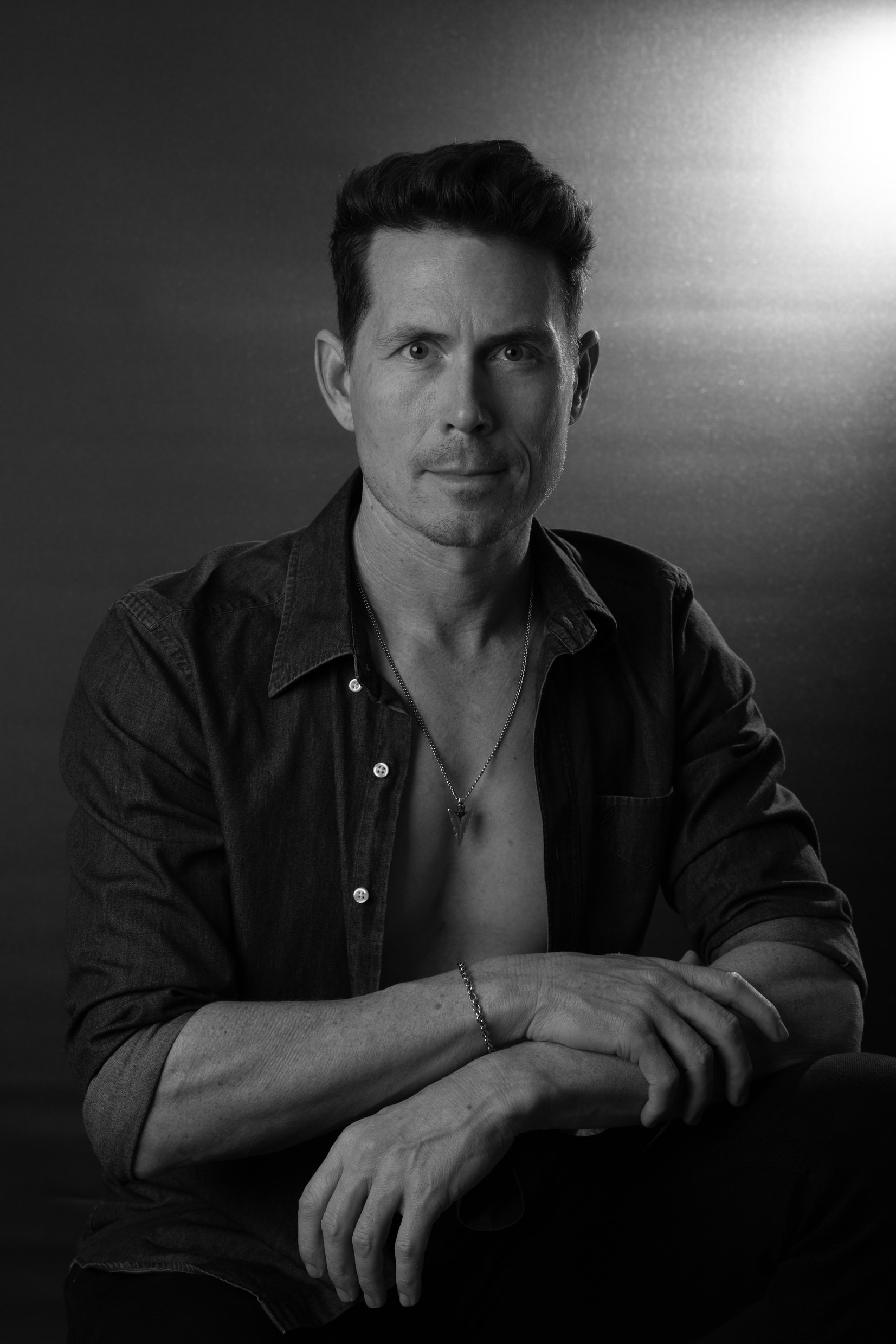 Dan Ormerod, seated with his arms folded, in an open dark shirt. A monochrome portrait lit from one side against a dark ground.